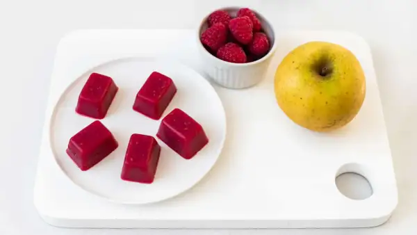 Cinq carrés de pâte de fruit rouge, des framboises et une pomme jaune sur une planche blanche. Une idée de conservation des fruits.